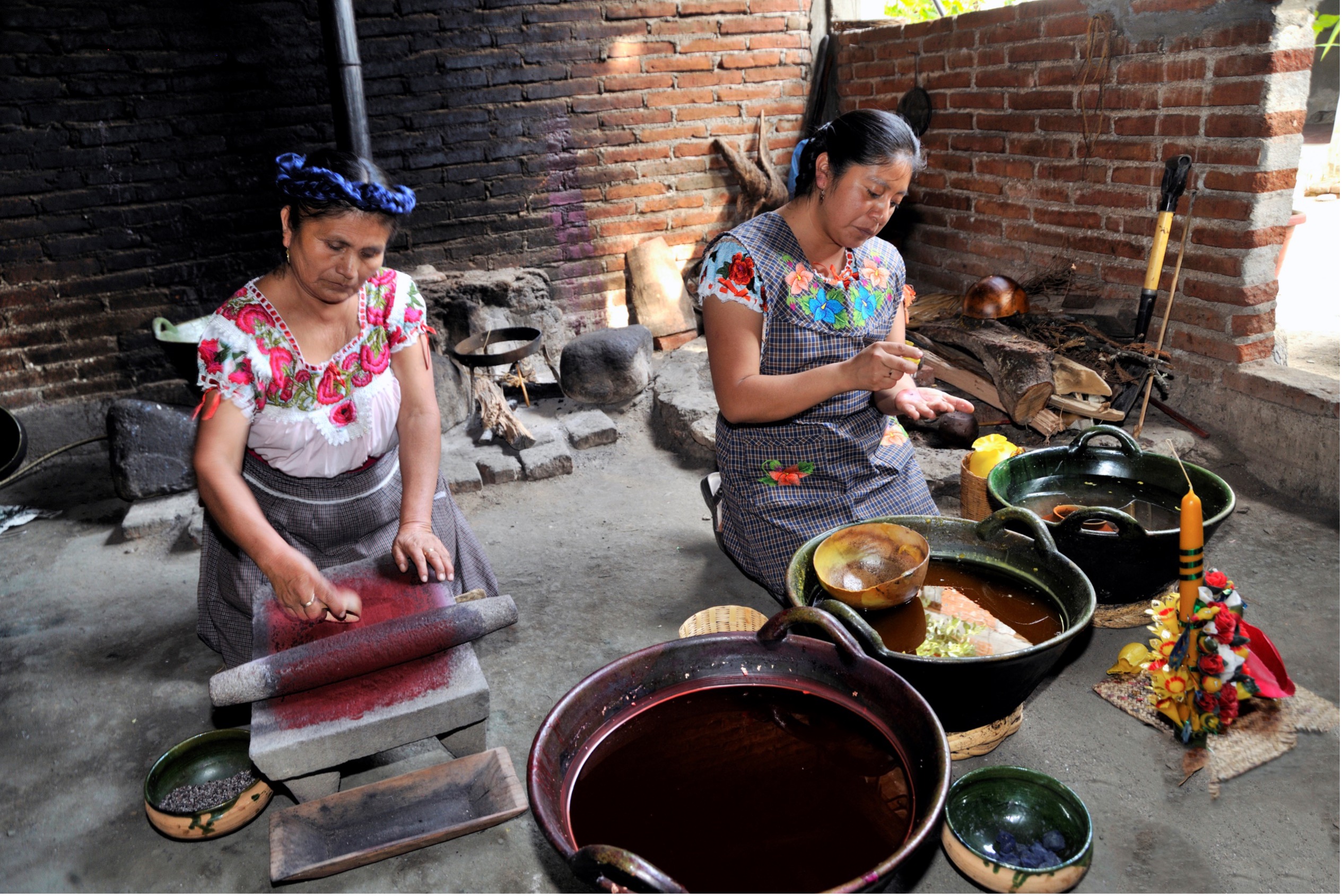 Two women sit side by side kneeling as they make candles. 