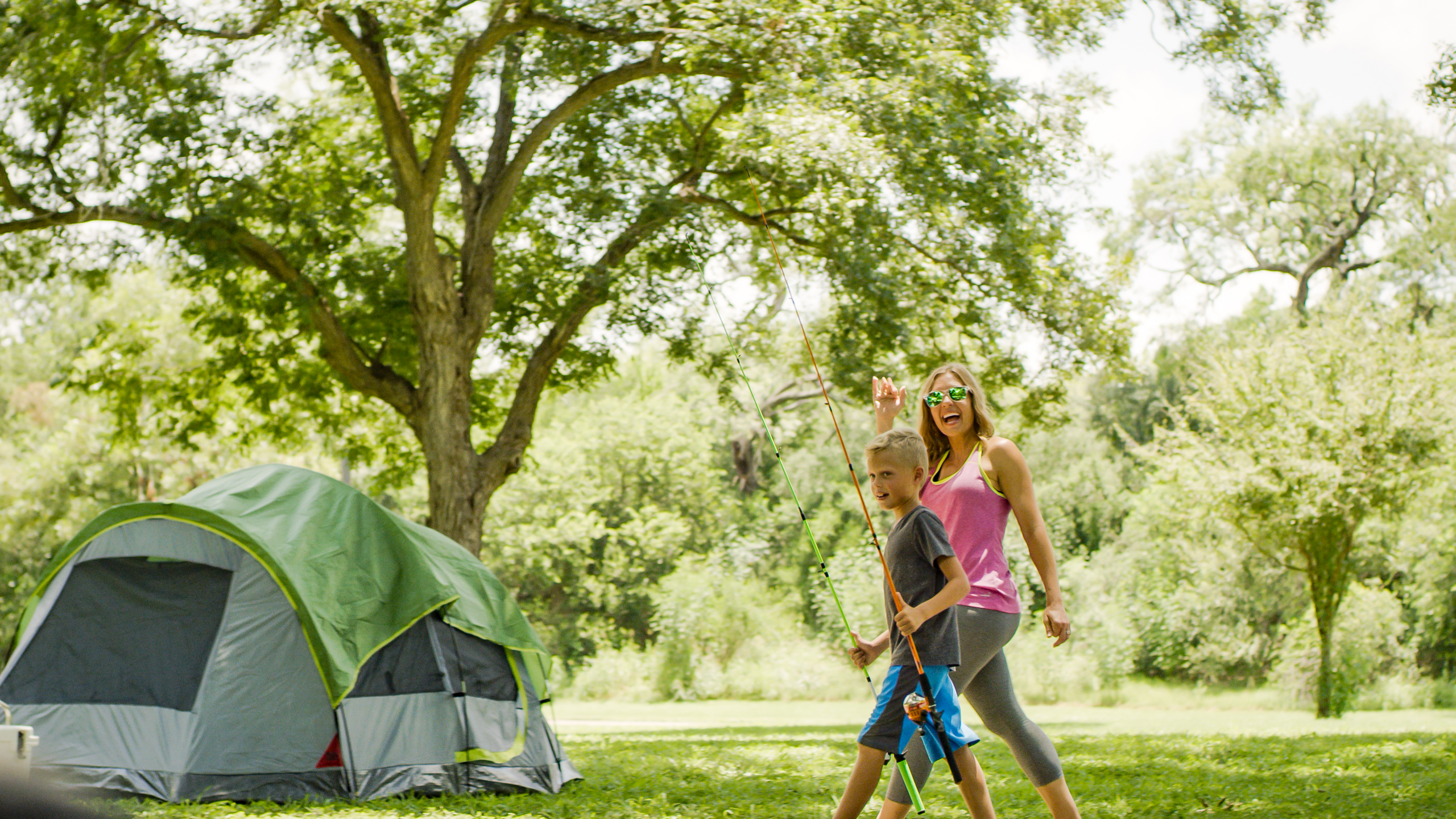 A mother waves as her child holds two fishing rods as they walk towards a camping site.