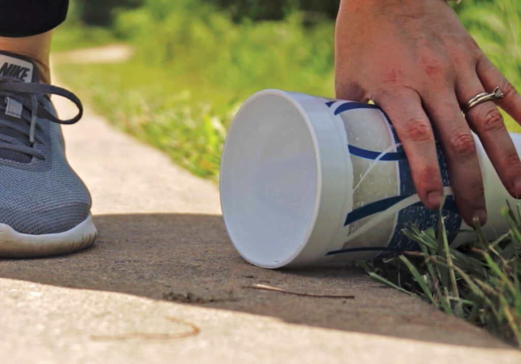 A hand reaches down to the ground to pick up a styrofoam cup.