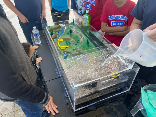 A water jug pours water into a watershed model demonstrating flooding patterns. 