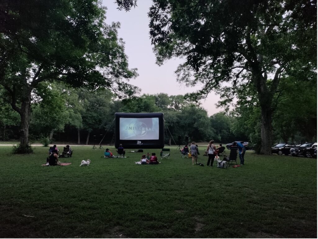 A large inflatable screens shows a movie in the middle of a park with families.