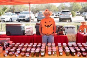 A man in a Halloween shirt and cowboy hat stands behind a table full of pickled items.
