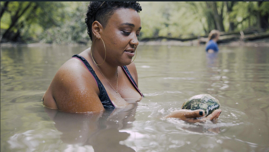 A woman holds a large mussel in her hands while in the river.