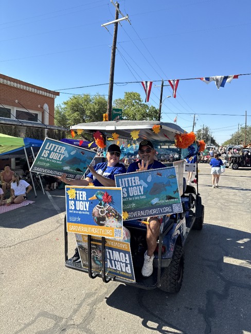 Two people sit in the back of a golf cart holding "Litter Is Ugly" signage